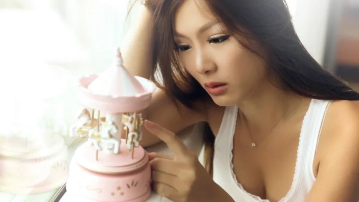 一名成年女子穿着白色无袖上衣，低头凝视手边的粉色旋转木马装饰。