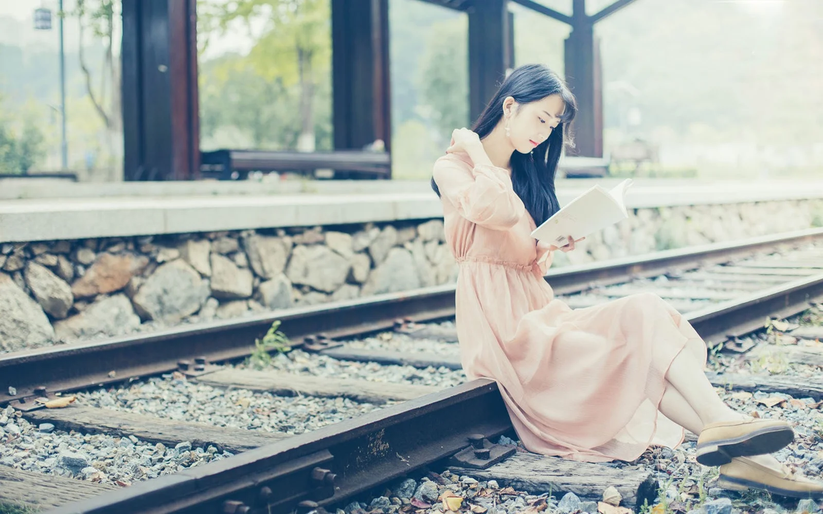 一名成年女子身穿浅色连衣裙坐在铁轨旁阅读书籍。