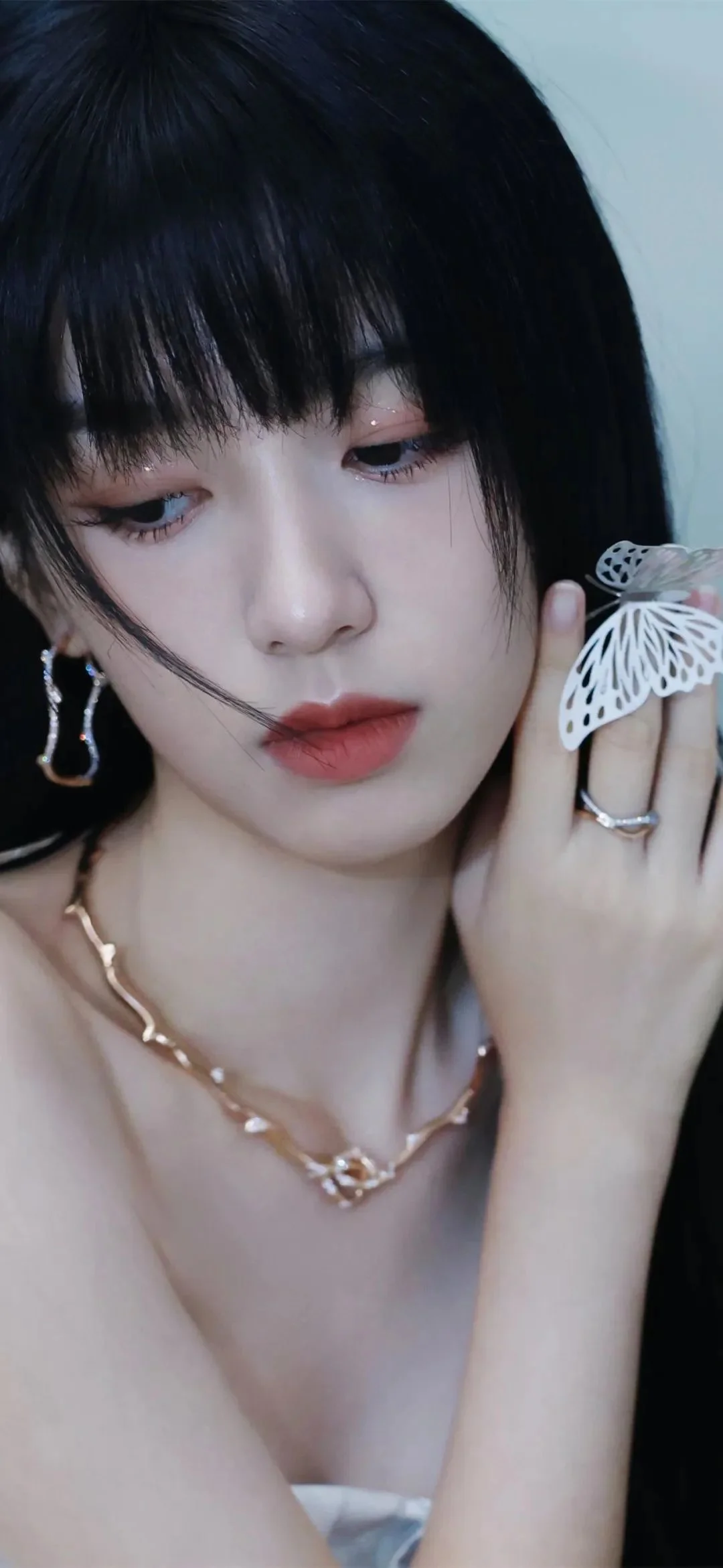 一位黑发女性近距离注视镜头，手持白色蝴蝶饰品，佩戴耳环和项链。