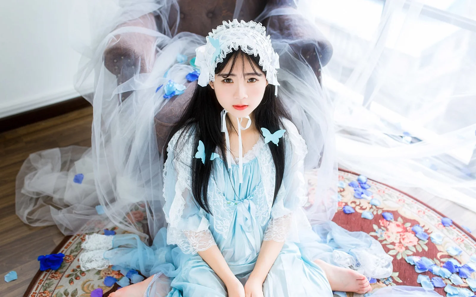 一名身穿浅蓝色服饰、戴白色头饰的女性坐在室内地面上，周围有半透明布料。