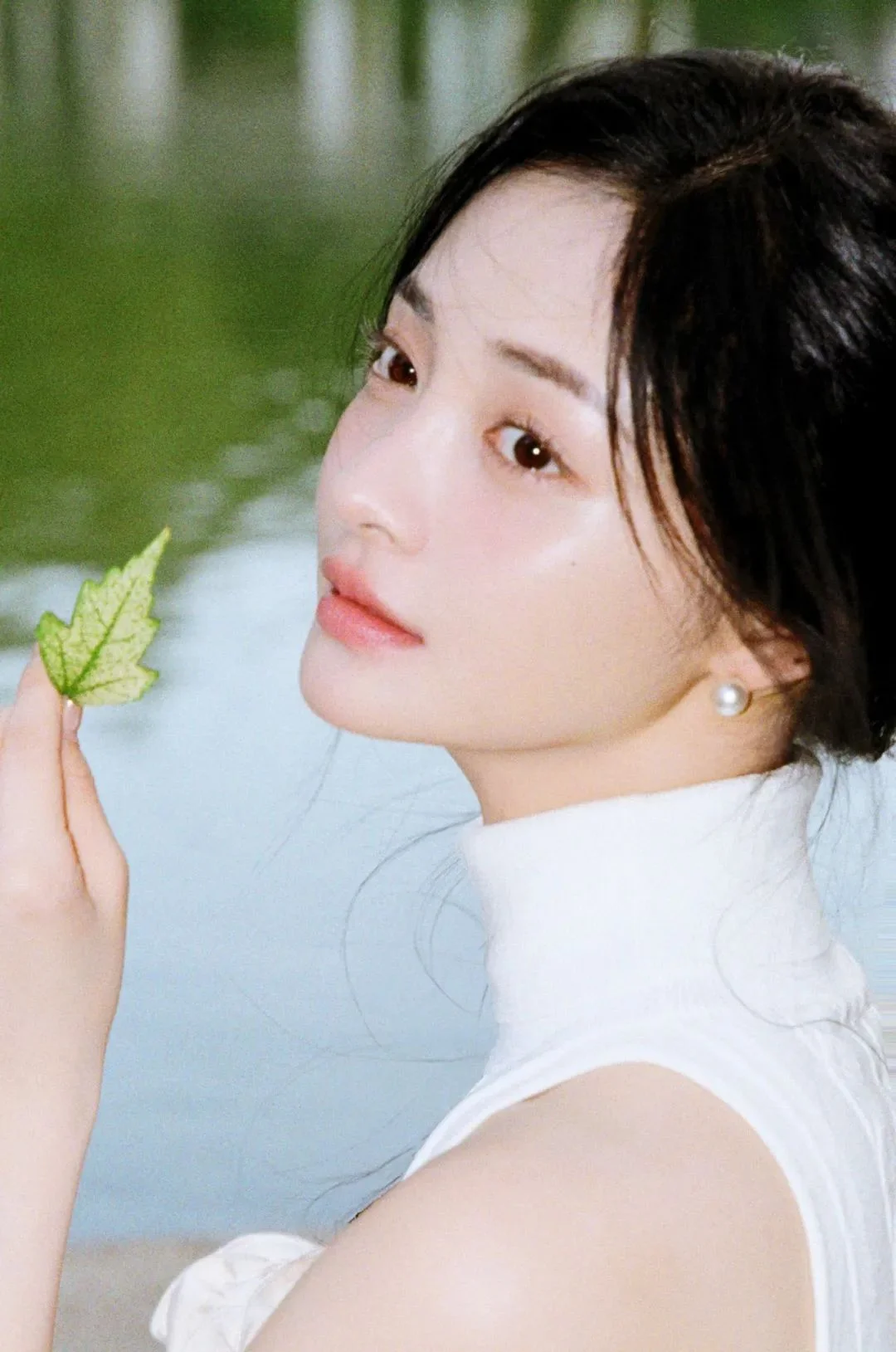 흰색 민소매 상의를 입은 여성이 부드러운 자연광 속에서 초록 잎을 들고 카메라를 바라보고 있다.
