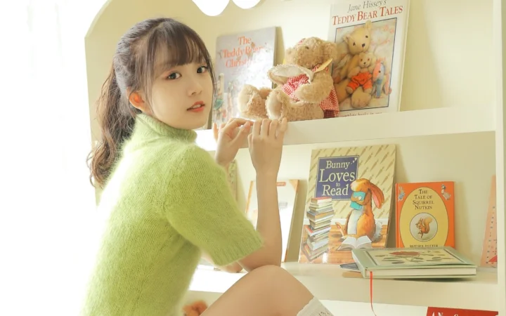 本やぬいぐるみが並ぶ本棚のそばに座った若い女性が、カメラを振り返っている。