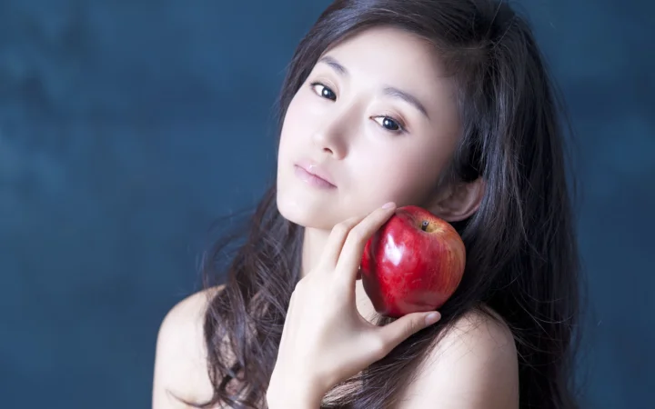 一名成年女性手持红苹果靠近脸颊，在蓝色背景前摆姿势。