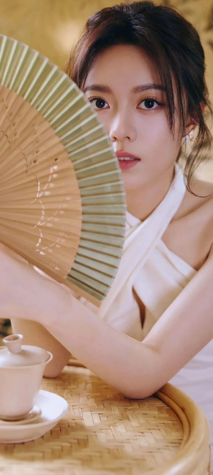 一名女子手持折扇坐在竹编桌旁，前景放着茶杯。
