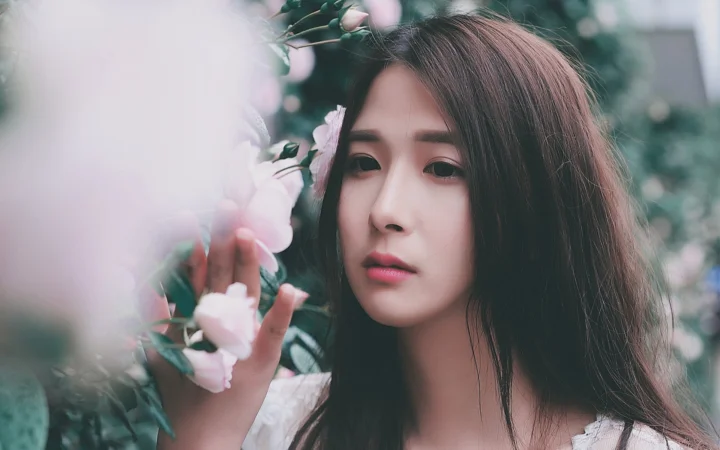 꽃과 푸른 잎사귀 사이에서 긴 머리 여성이 카메라를 바라보고 있다.