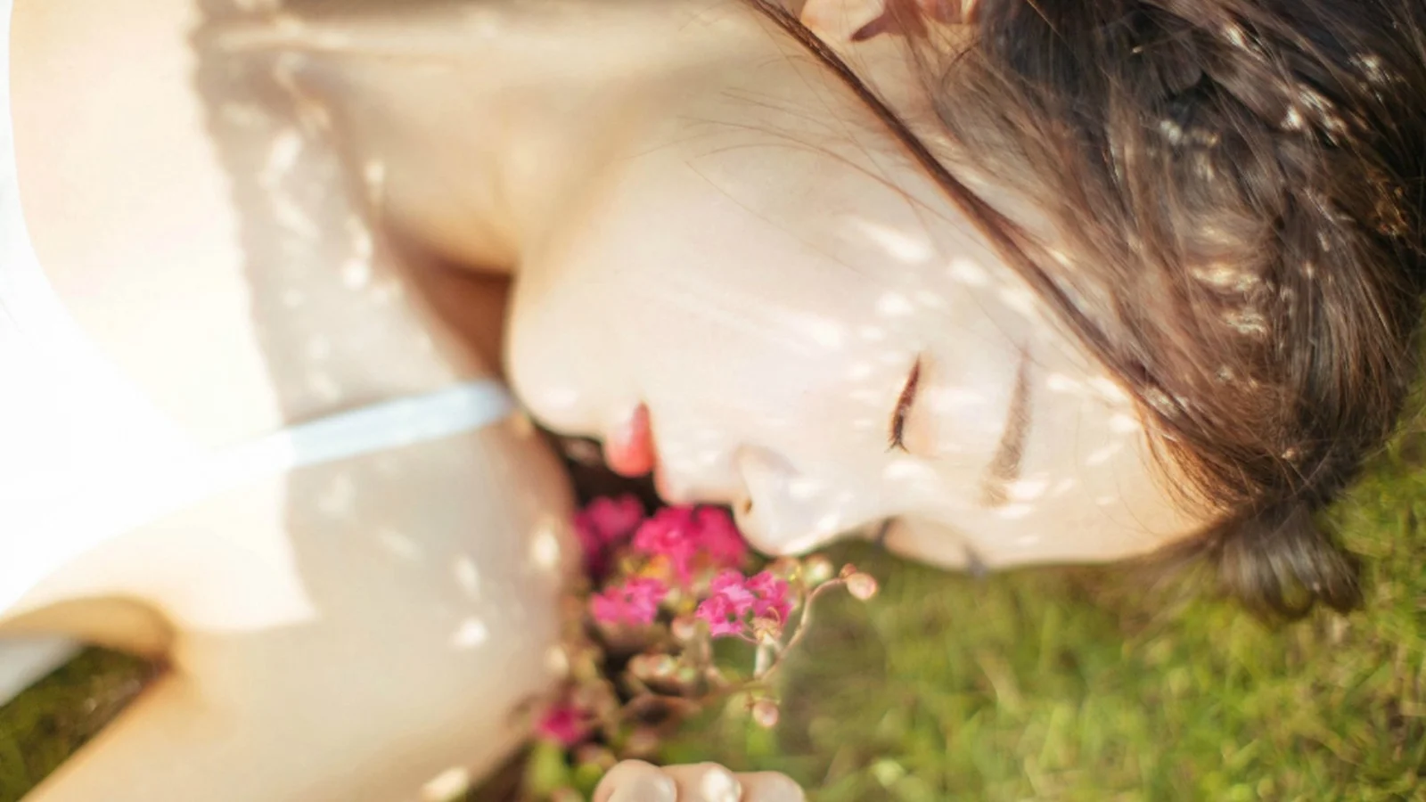 一名女性侧脸闭眼躺在草地上，脸旁有一束粉色小花。