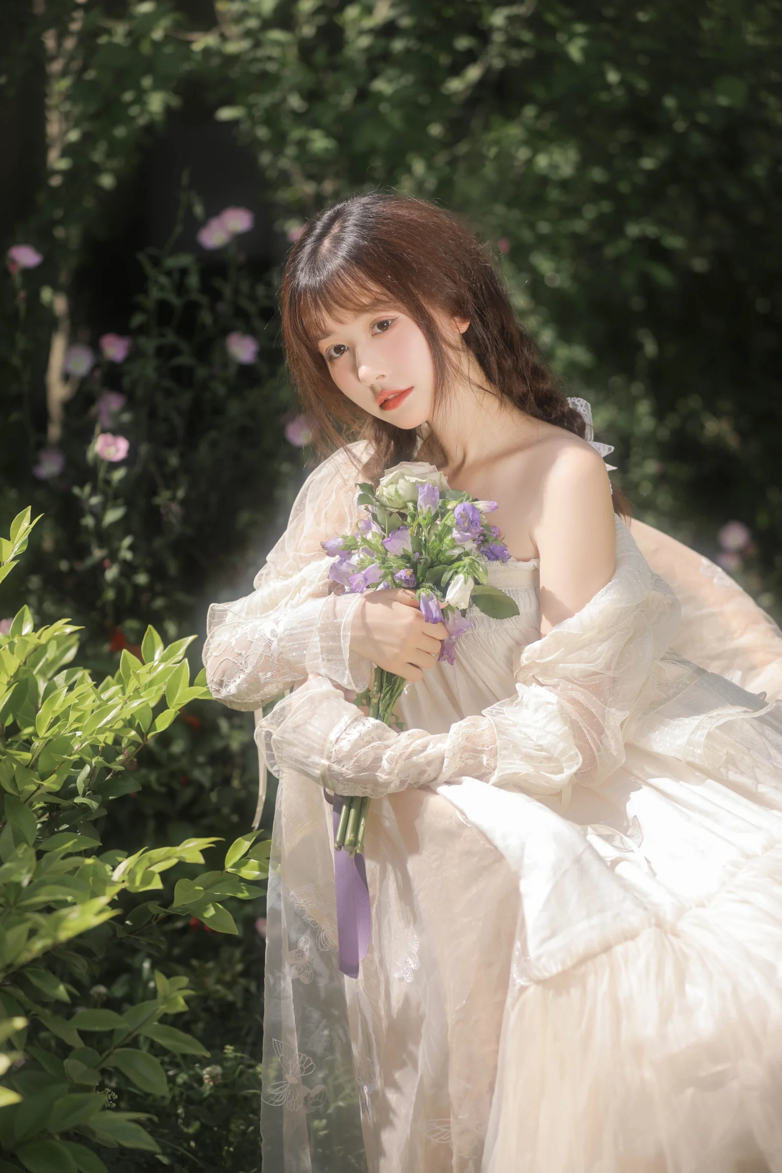 흰 드레스를 입은 성인 여성이 초록이 우거진 정원에서 꽃다발을 들고 있다.