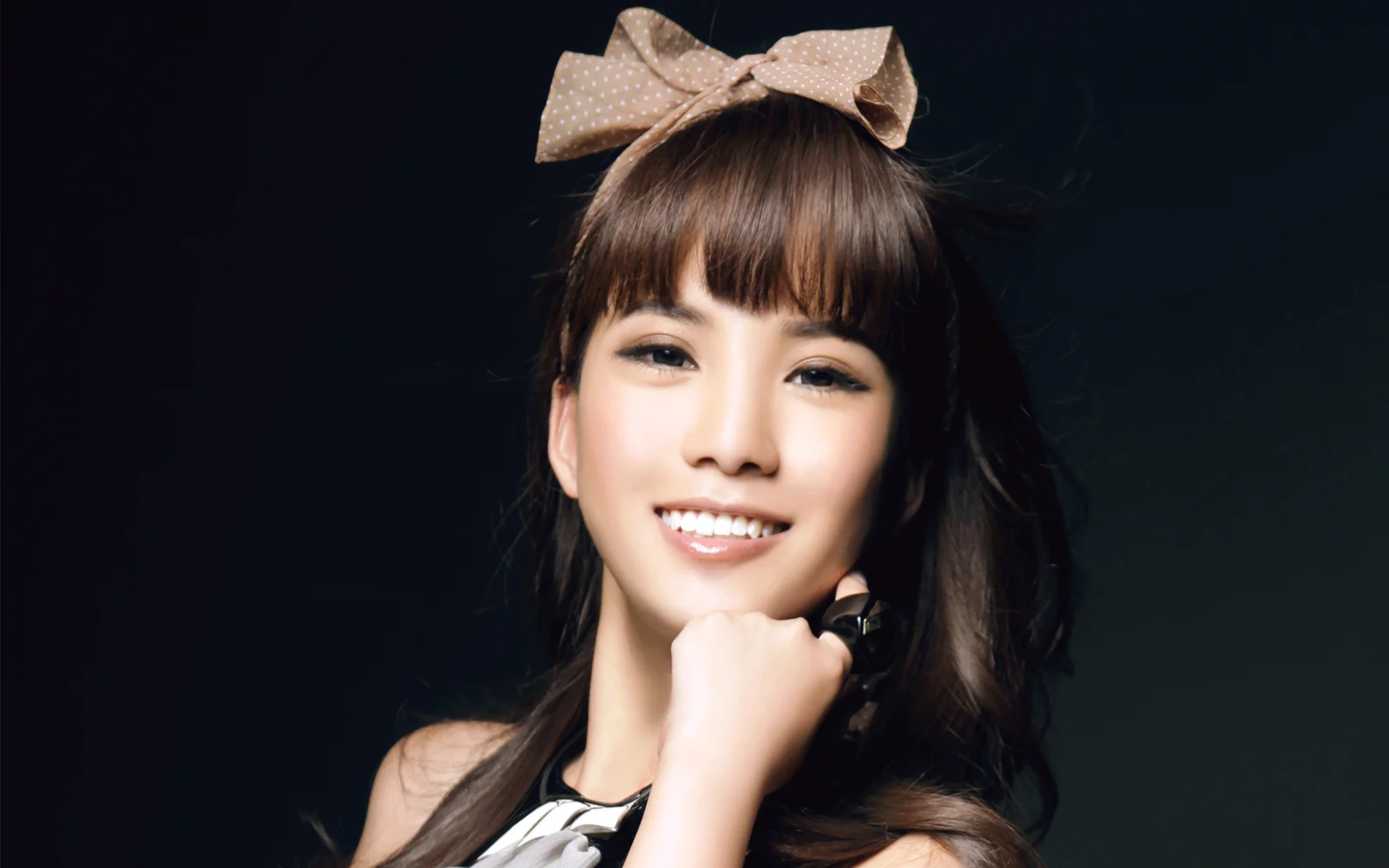 어두운 배경 앞에서 리본 머리 장식을 한 여성이 카메라를 바라보며 미소 짓고 있다.