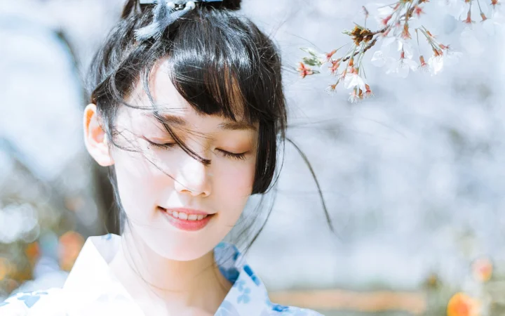 검은 머리의 여성이 눈을 감고 미소 지으며 부드러운 벚꽃과 밝은 야외 배경 앞에 서 있습니다.