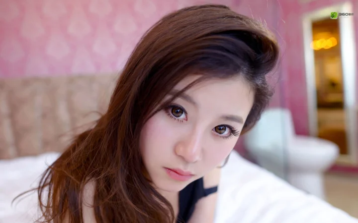 一名棕发女性近距离面对镜头，背景是柔和的粉色室内环境。