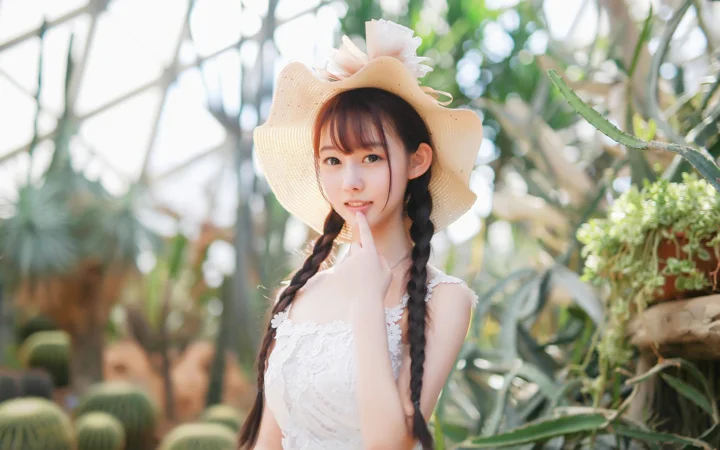 一名戴着宽檐帽、编着双辫的女子站在阳光明亮的温室植物间。