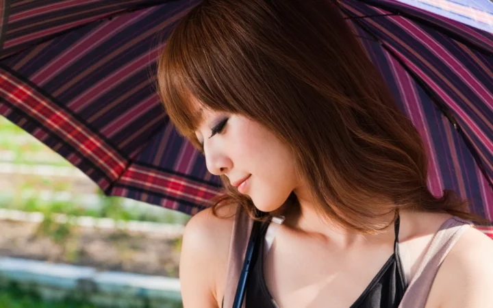 一名棕发女子在彩色条纹伞下低头侧望，背景隐约可见户外绿意。