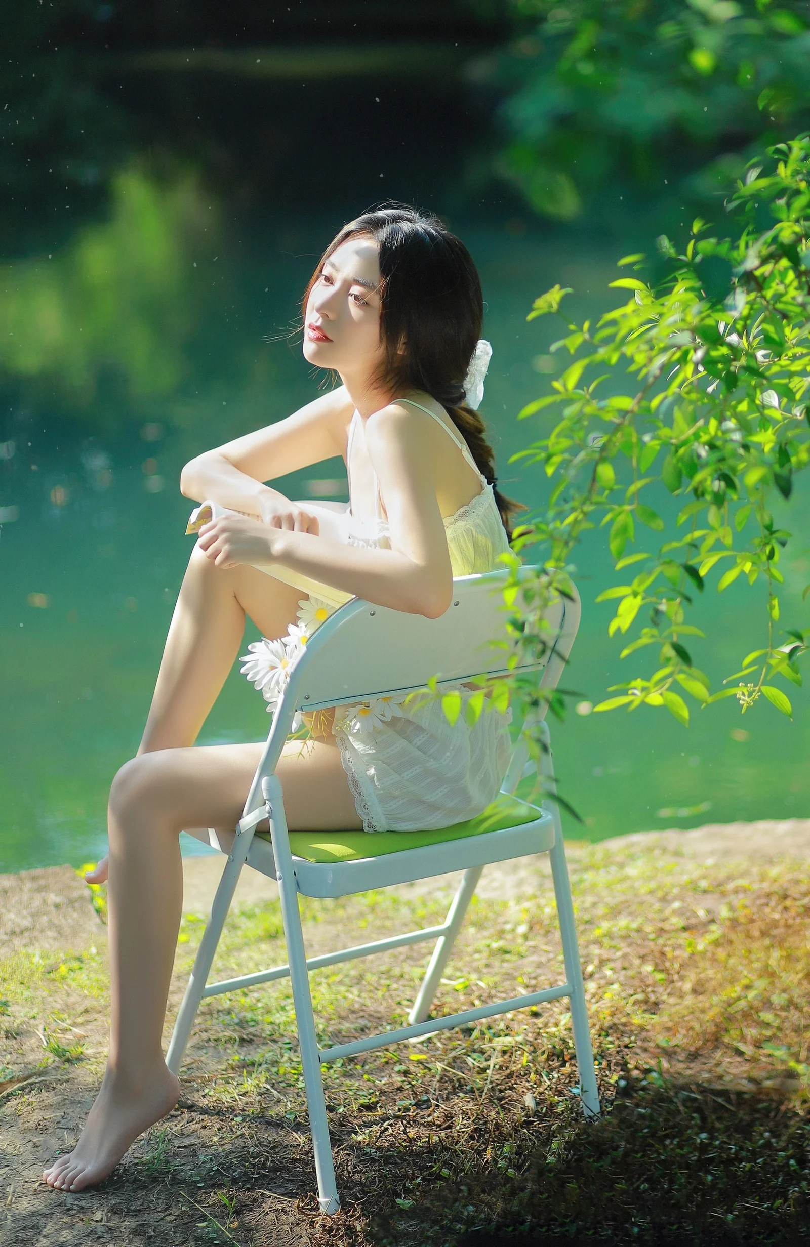 흰색 원피스를 입은 여성이 초록빛 물과 나무 곁의 접이식 의자에 앉아 있다.