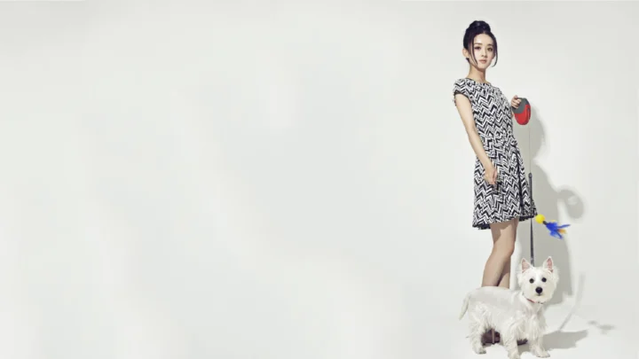 흑백 무늬 원피스를 입은 여성이 흰색 배경 앞에서 흰 개와 함께 서 있다.