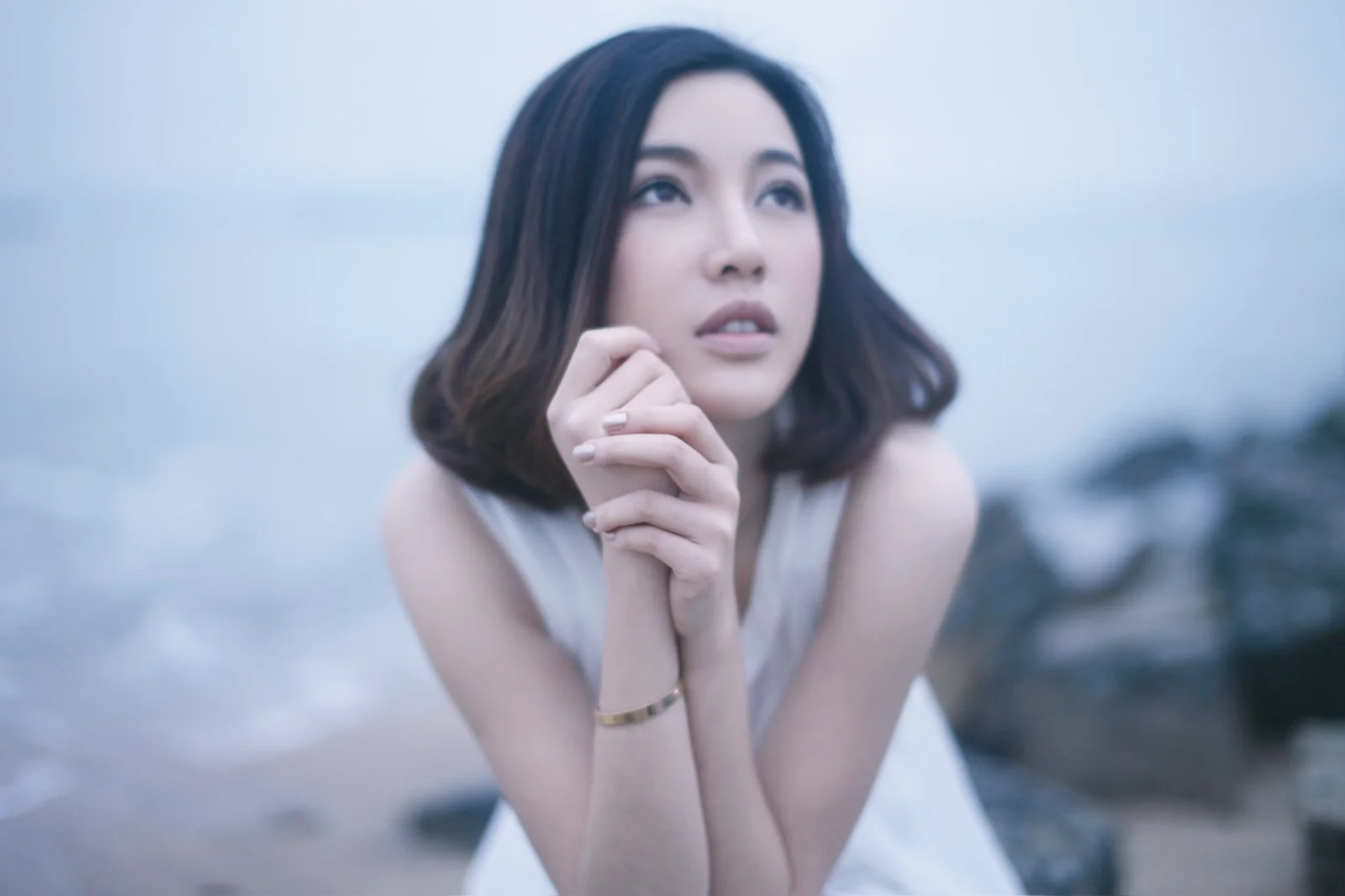成人の女性が手を組み、柔らかな青い色調の屋外背景の前で遠くを見つめている。