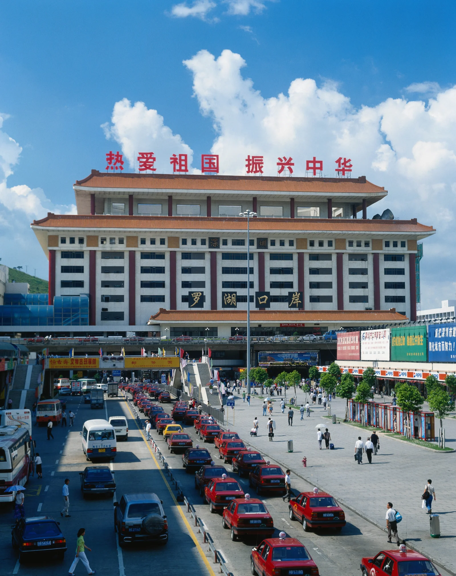 一座带有红色标语的大型城市建筑矗立在蓝天白云下，前方街道上车辆和行人来往。