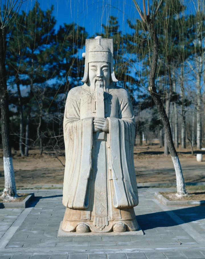 一尊身着长袍、手持法器的石雕人物立在树木环绕的石板庭院中。