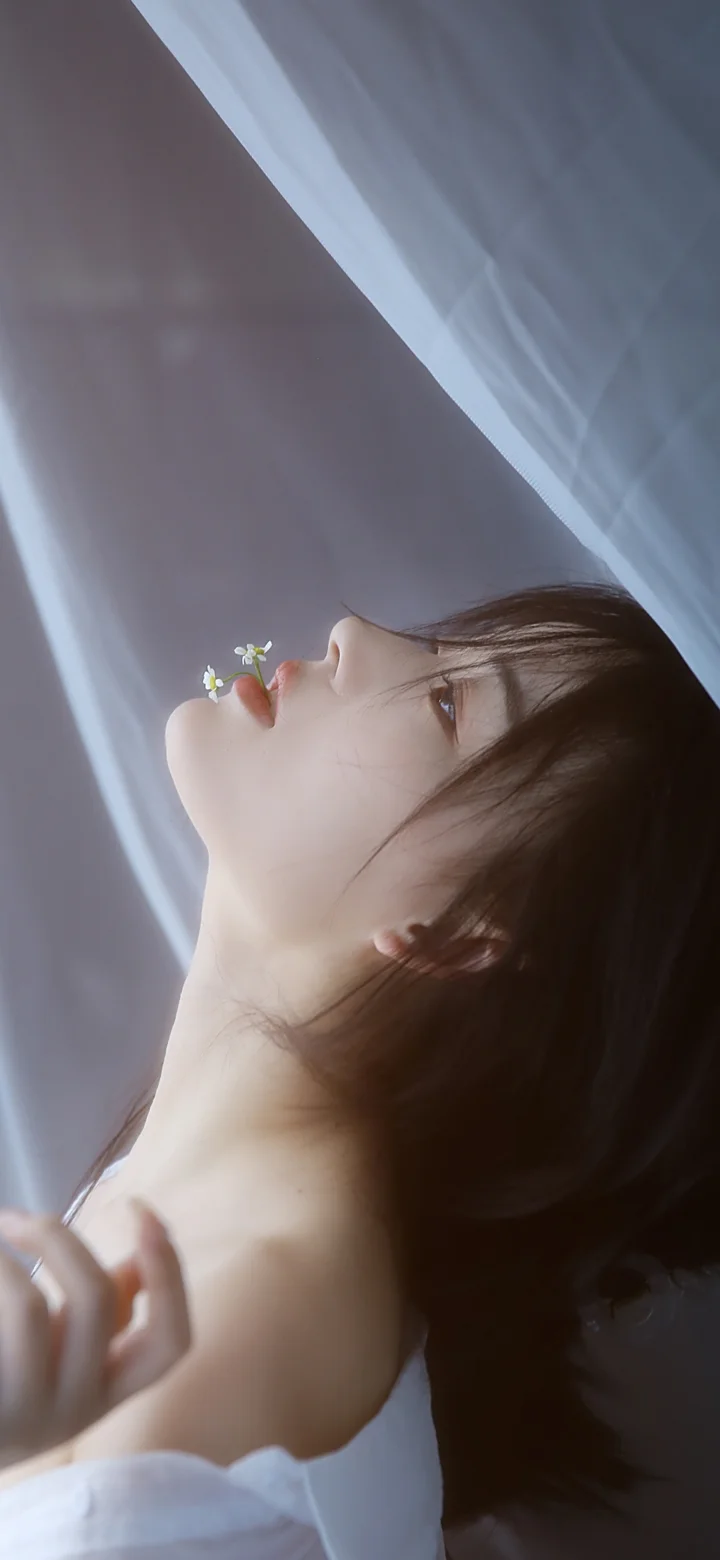 긴 머리의 여성이 작은 꽃 가까이에 얼굴을 대고 흰색 옷을 입은 채 부드러운 청회색 배경에 담겨 있다.