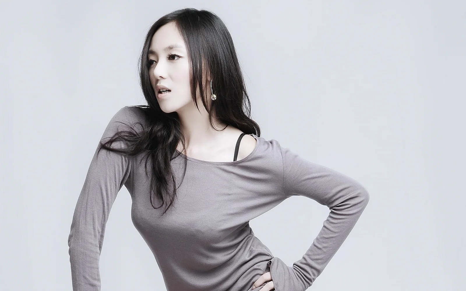 一名长发女性穿着灰色长袖上衣，在浅灰色背景前侧身摆 pose。