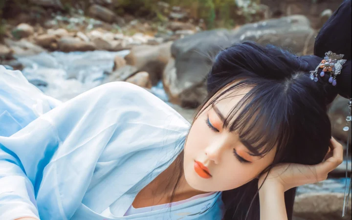 一名身穿浅蓝色古风服饰的女子在溪边侧卧，周围可见岩石与流水。