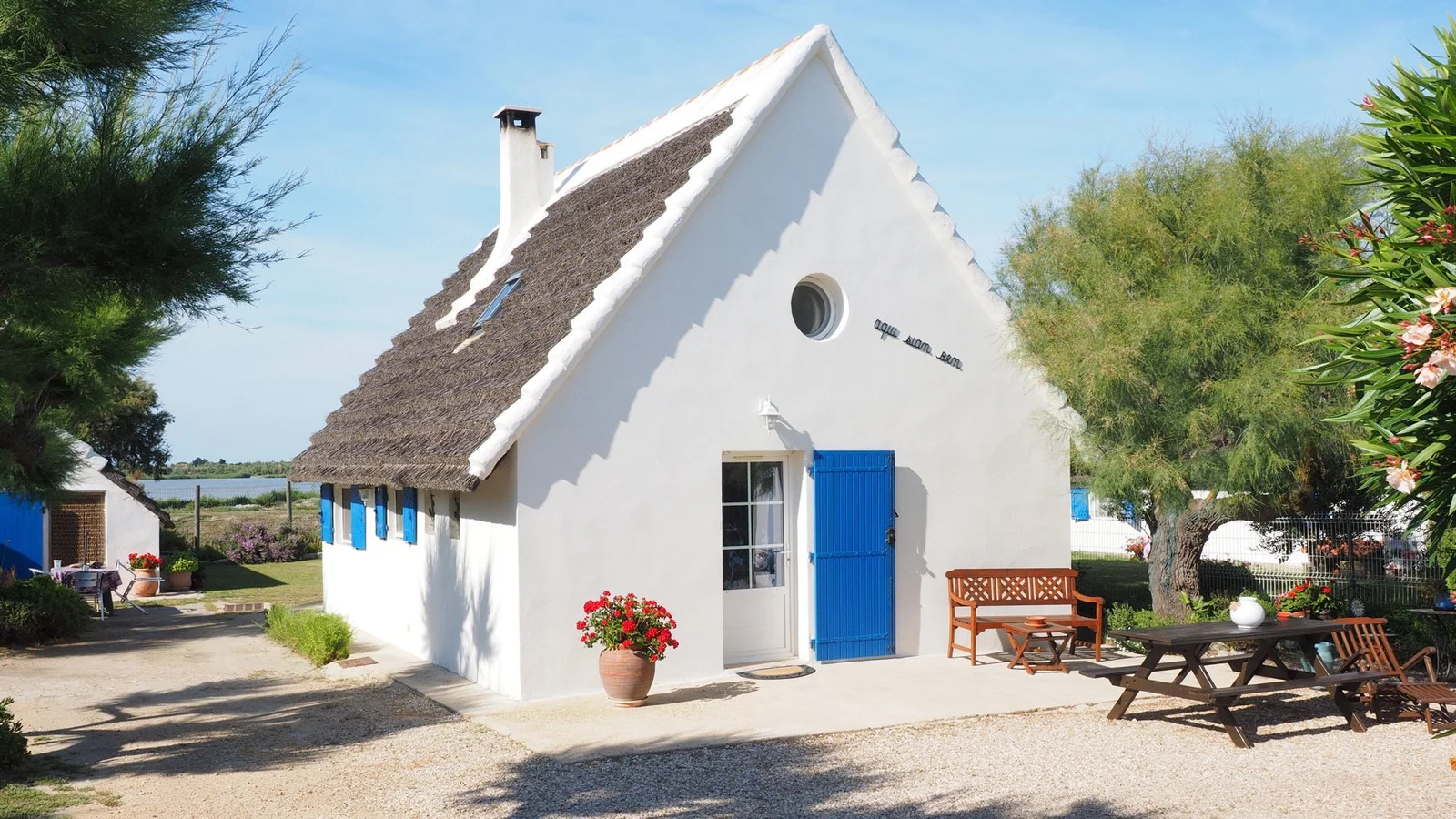 日差しの中、青い扉と窓のある白い茅葺き cottage が庭に建ち、そばにベンチとピクニックテーブルが置かれています。