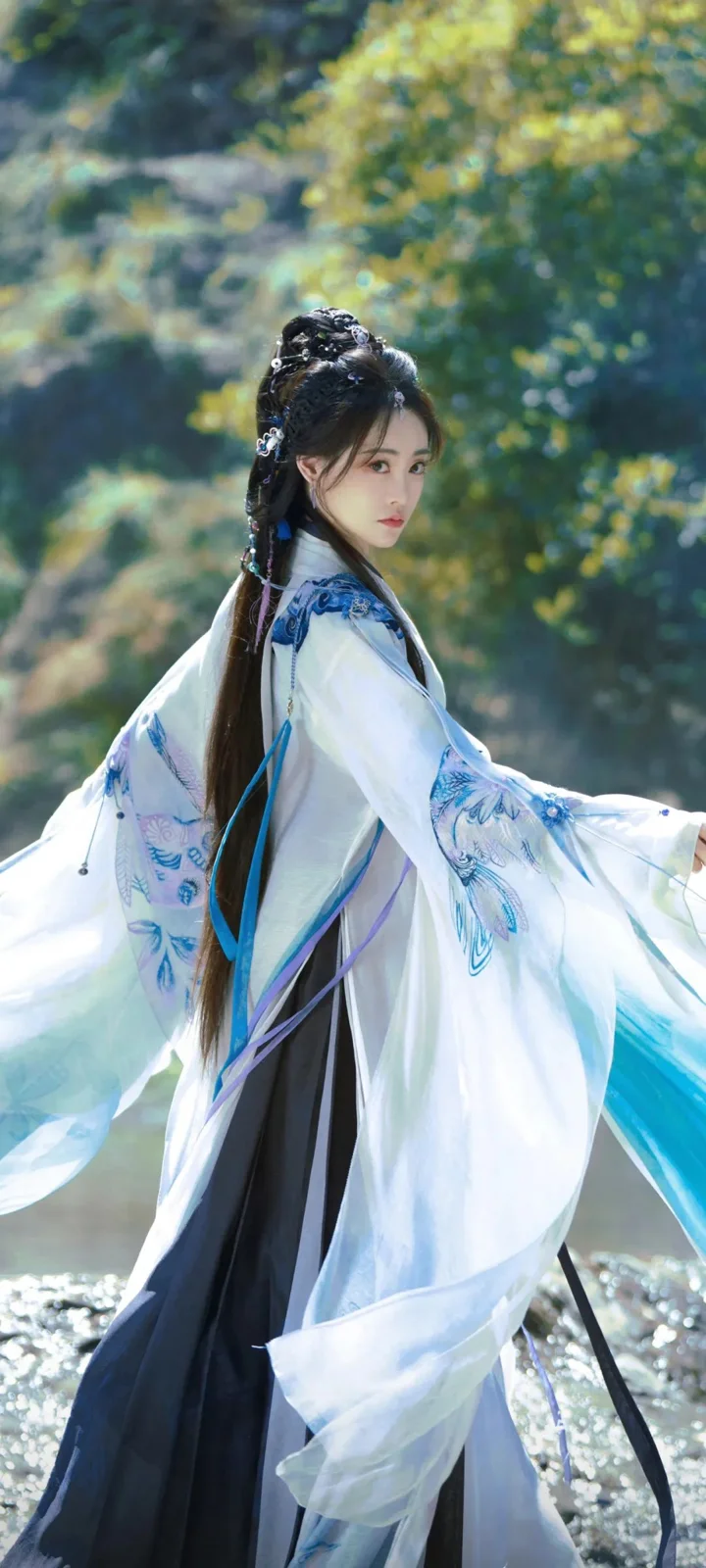 一名身穿白蓝色传统服饰的女子站在阳光斑驳的林间。
