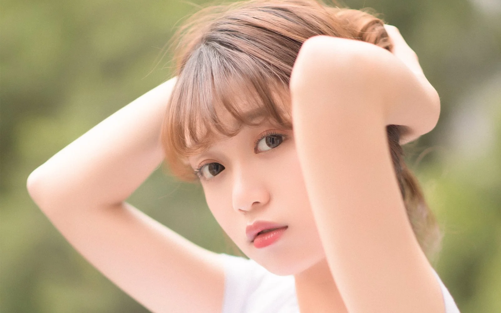 갈색 머리의 여성이 부드럽게 흐린 야외 배경 앞에서 머리를 정리하고 있습니다.