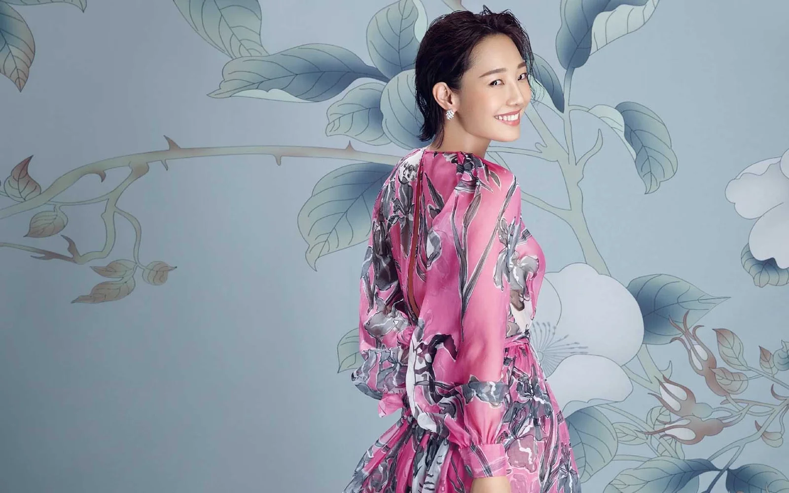 一名身穿粉色花卉图案服装的成年女性站在淡蓝色花卉背景前微笑回望。