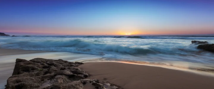 夕阳映照下的海浪涌向沙滩，前景散布着深色礁石。