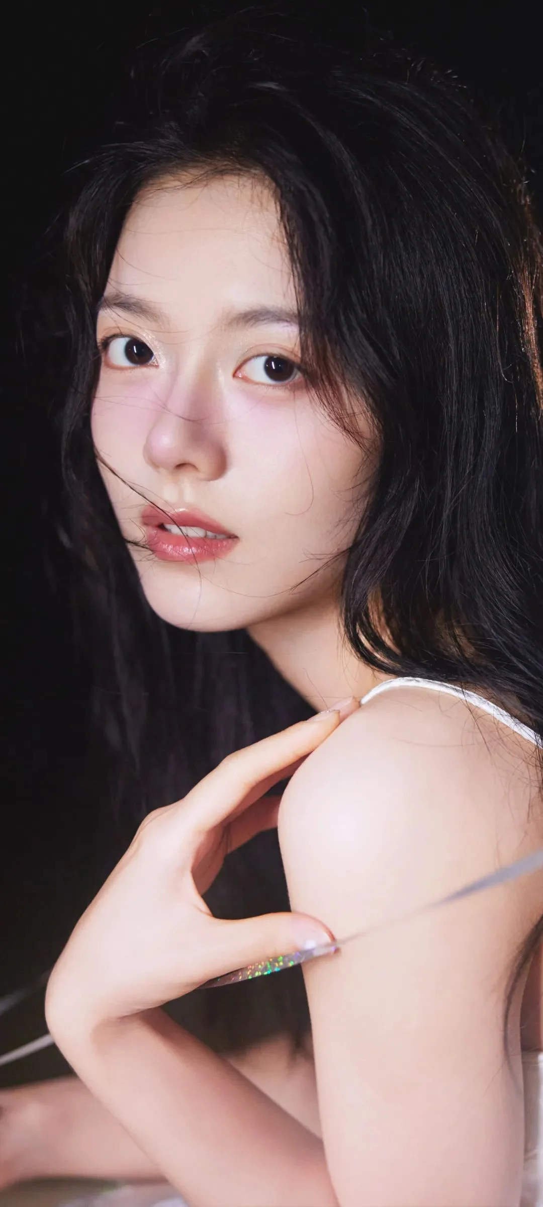 黒髪の女性が暗い背景の前で横顔を見せながらカメラを見つめ、腕と肩が写っている。