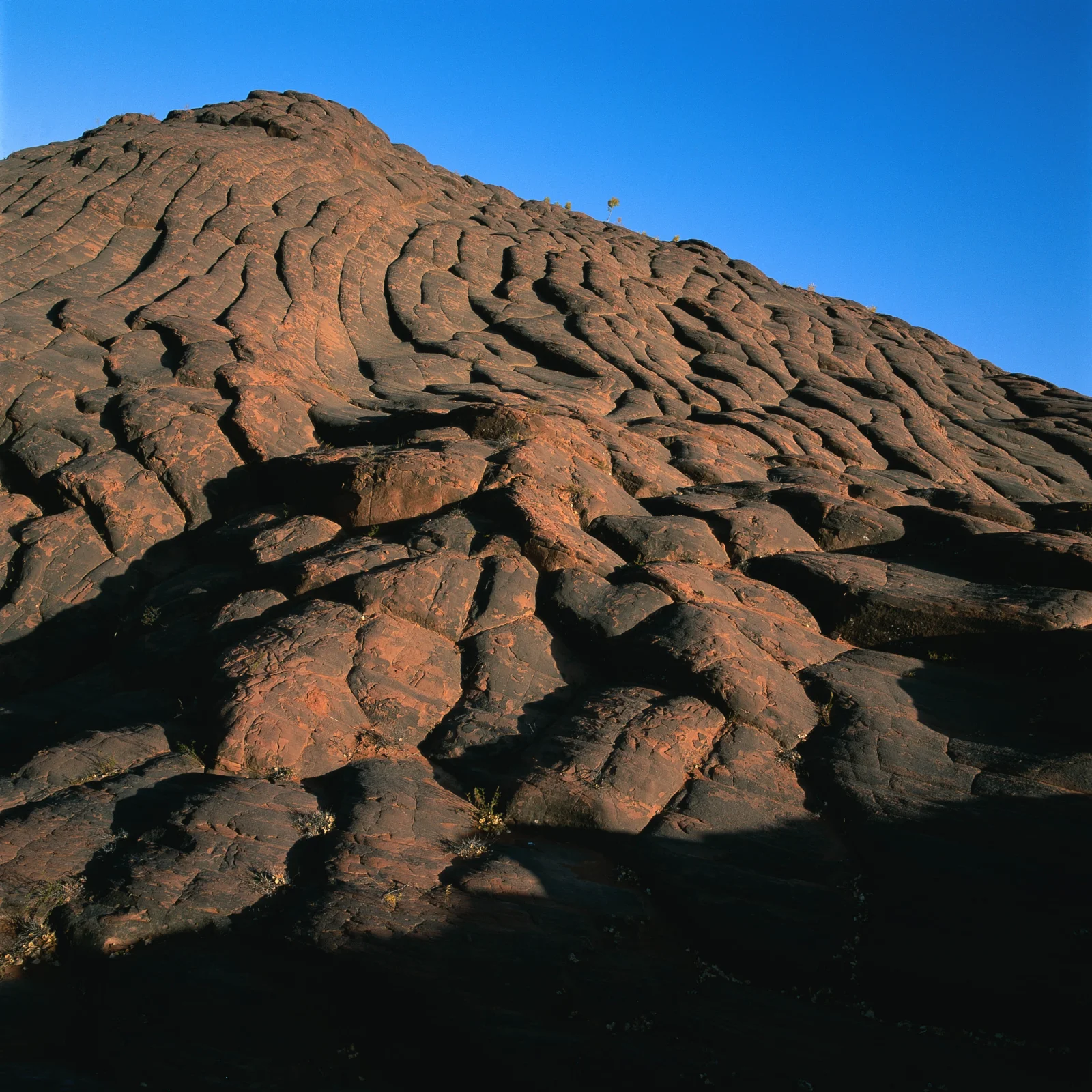 阳光照亮了蓝天下布满沟壑的深色火山岩山坡。