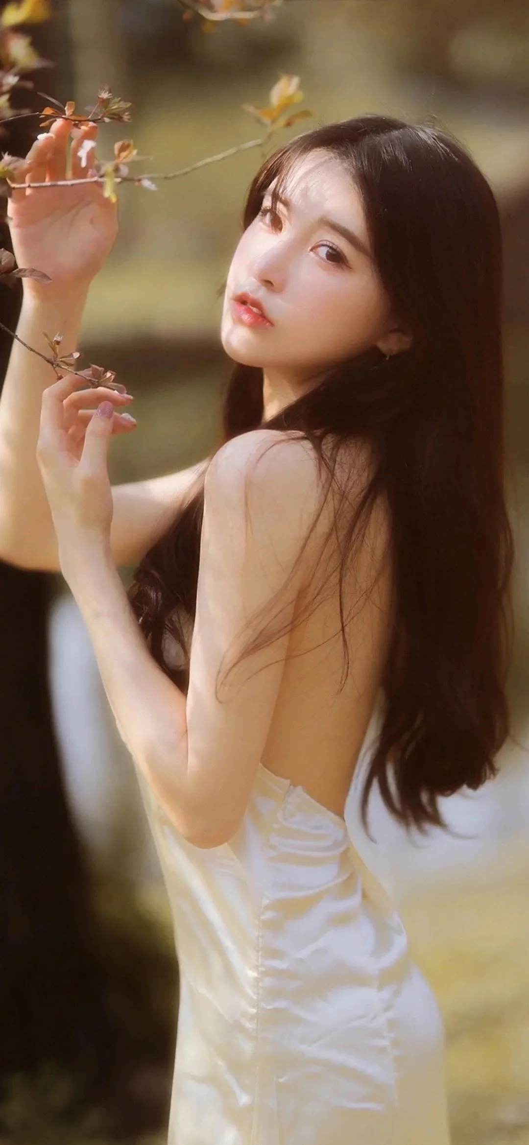 긴 머리의 여성이 부드러운 숲의 빛 속에서 나뭇가지 곁에 서서 카메라를 돌아보고 있다.