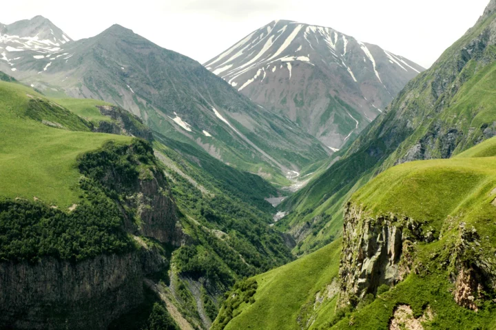 绿色山坡环绕着深邃山谷，远处耸立着带有积雪的群山，谷底可见蜿蜒溪流。
