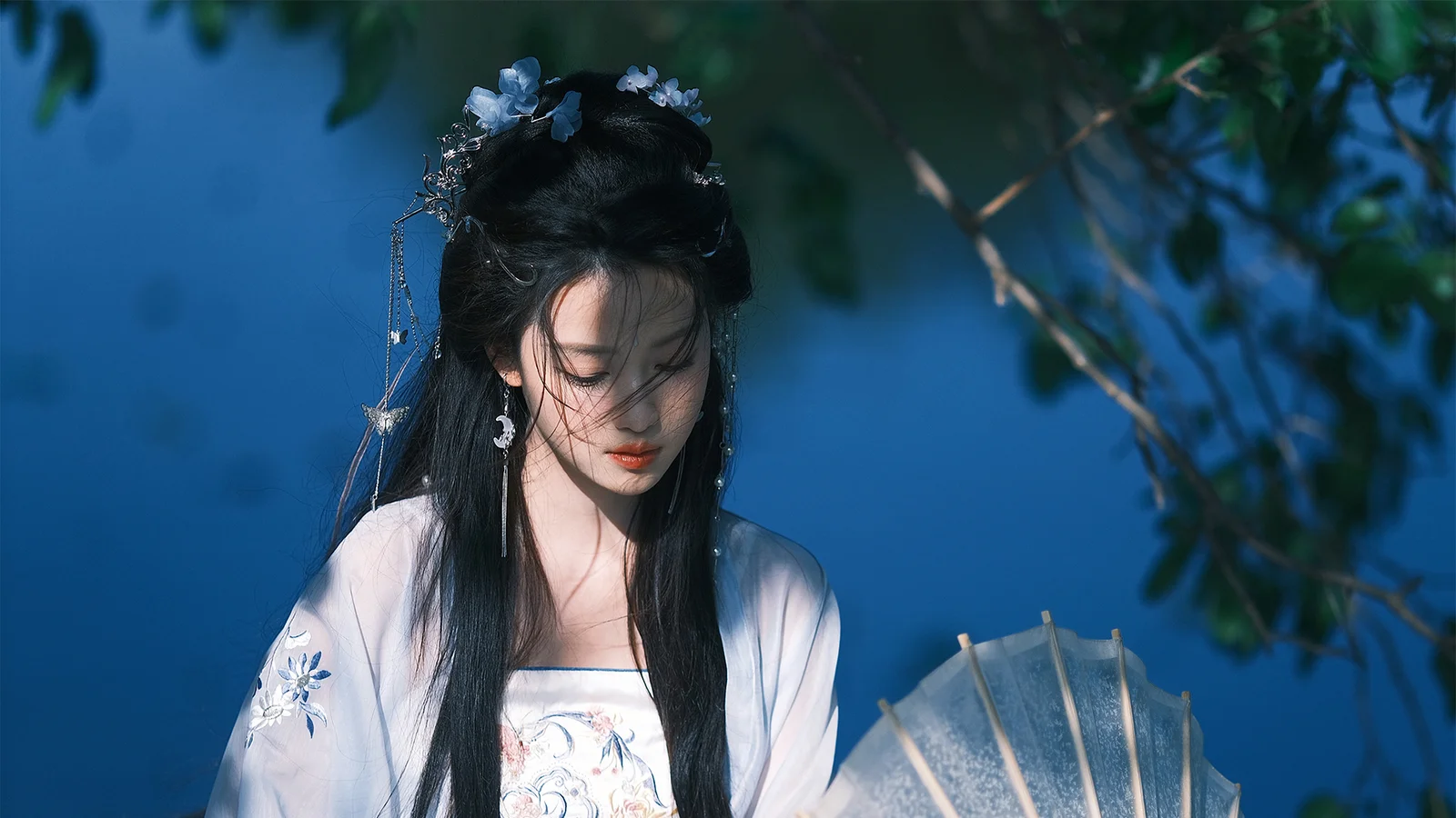 전통 의상을 입은 성인 여성이 반투명한 양산을 들고 푸른 배경 앞에 서 있으며 주변에 나뭇잎이 보입니다.