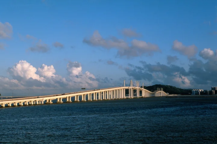 一座白色长桥横跨深蓝色水面，远处是低矮山丘与建筑，天空中点缀着云朵。