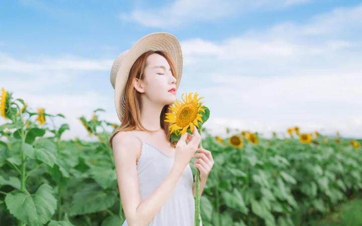 一名戴草帽的女子站在向日葵田中，手持一朵向日葵，背景是蓝天白云。