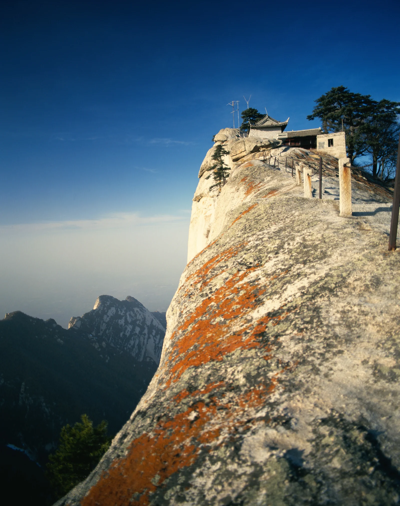 险峻的岩石山崖上坐落着一座古寺，远处群峰笼罩在湛蓝天空下。