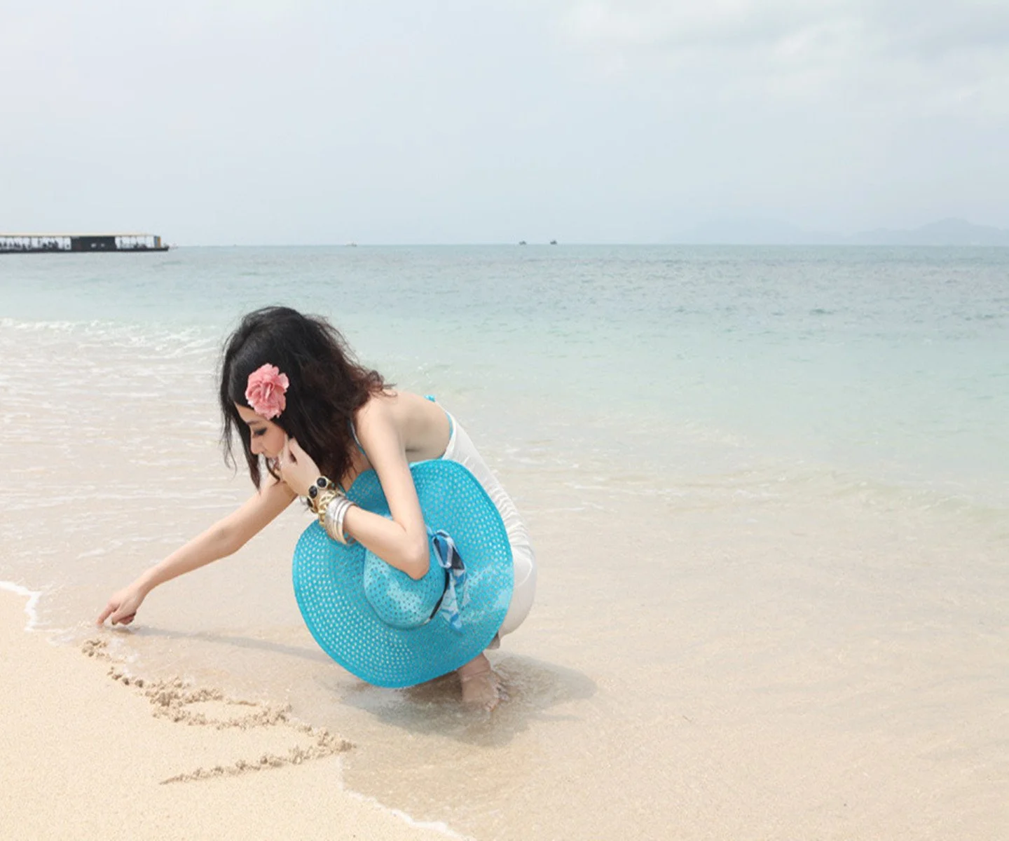 一名女子蹲在浅水沙滩上用手触碰沙面，身旁放着蓝色浮圈，远处是平静的海面。