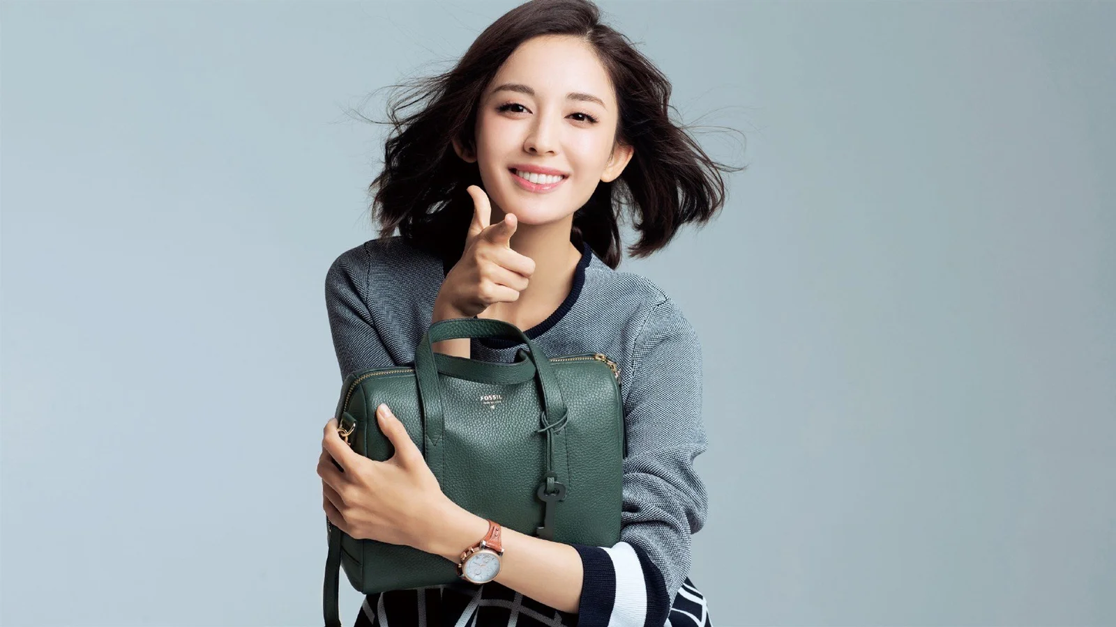 연한 회청색 배경 앞에서 성인 여성이 짙은 녹색 핸드백을 안고 미소 짓고 있습니다.