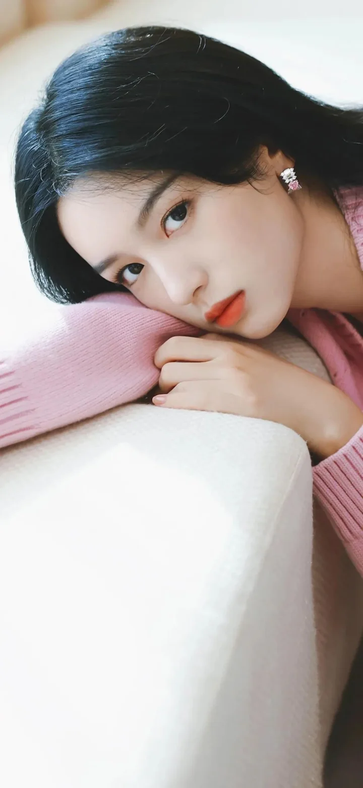 一位成年女性穿着粉色毛衣，侧躺在柔软的浅色表面上看向镜头。