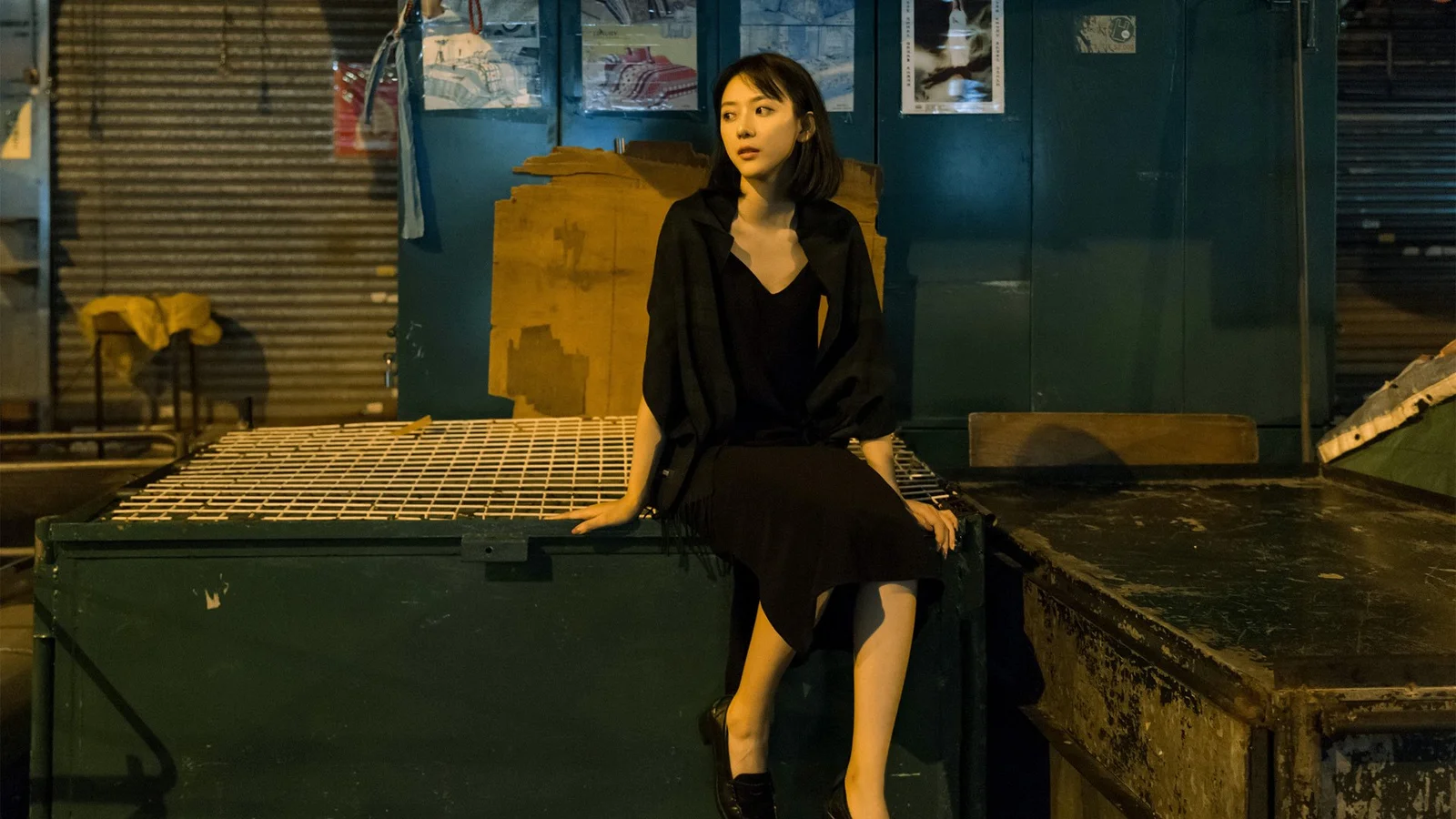 一名身穿黑色连衣裙的女子坐在昏暗城市环境中的金属平台上。