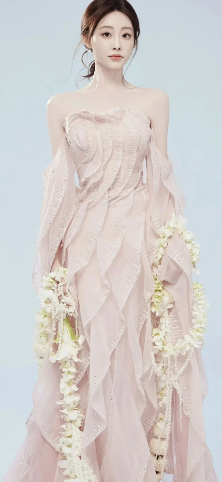 淡い青色の背景の前で、ピンクのロングドレスを着た成人女性が白い花飾りを手にしています。
