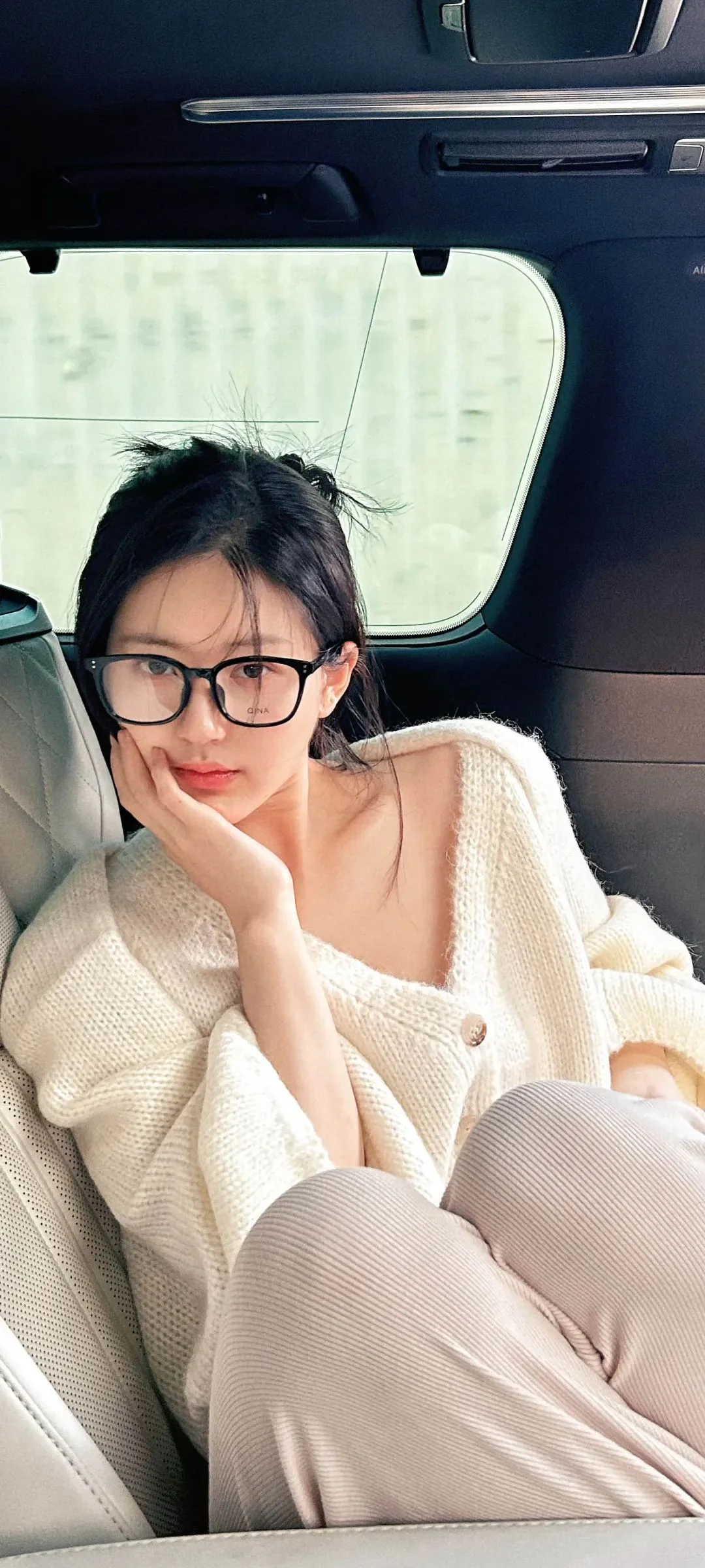 一名戴眼镜的女性坐在车内，身穿浅色针织衫和米色长裤，手托着下巴。
