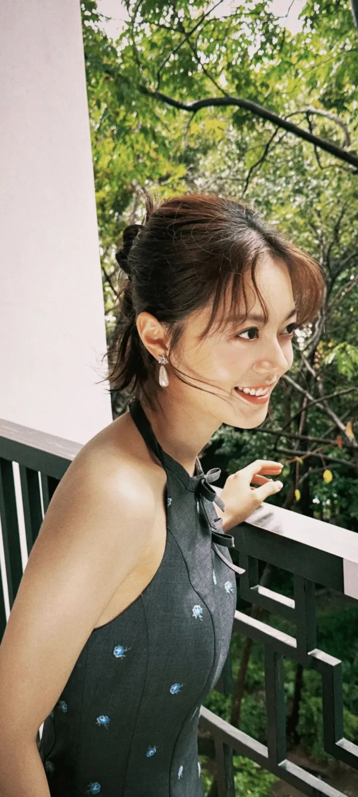 女性がベランダの手すりのそばに立ち、背後に緑の木々が広がっています。