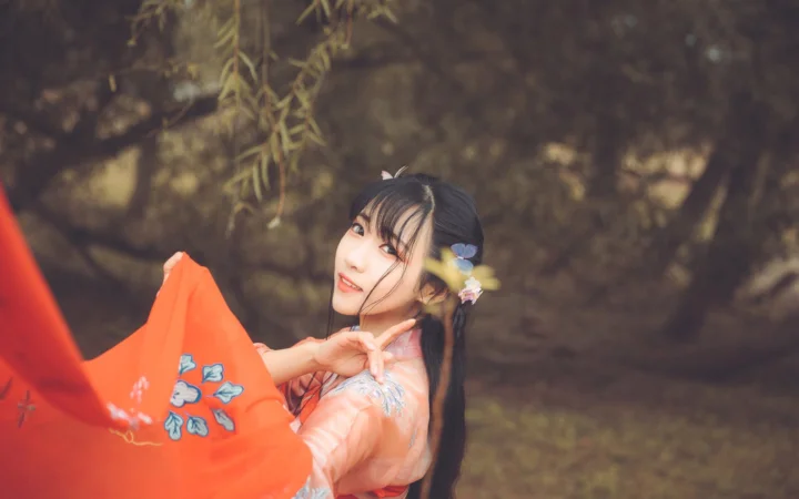 一名身穿橙色傳統服飾的女性在樹木背景前側身微笑。