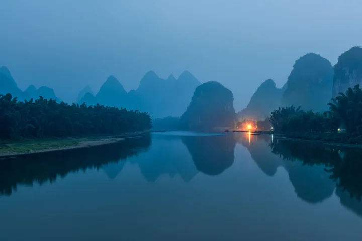 清晨薄雾笼罩着群山与漓江，远处灯火倒映在平静的水面上。
