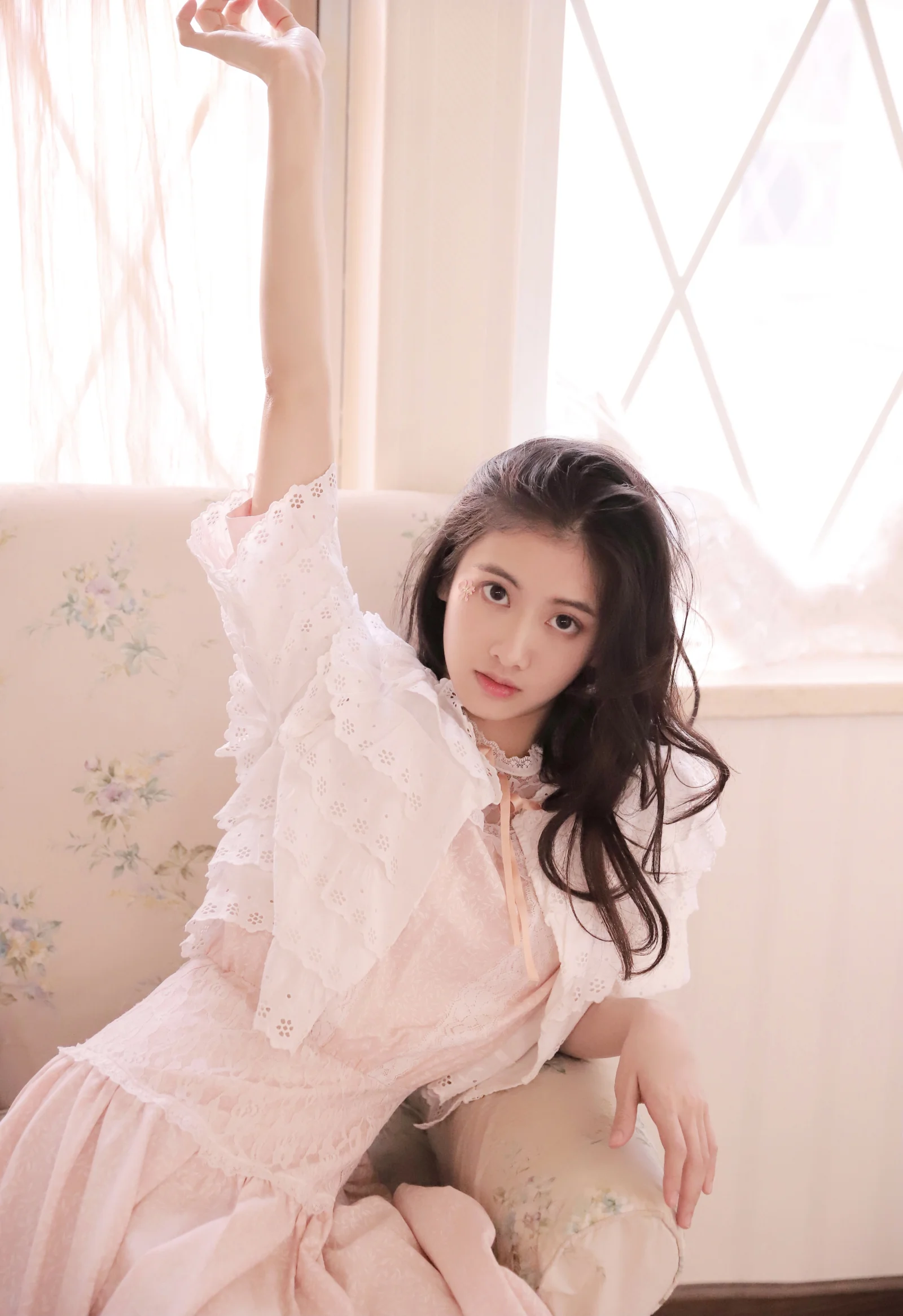 淡いピンクのドレスを着た女性が室内の窓辺に座り、片脚を上げてカメラを見つめています。