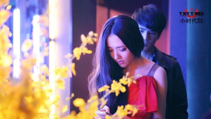 一名身穿红裙的女性站在一名男性前方，周围可见黄色花朵和紫蓝色灯光。