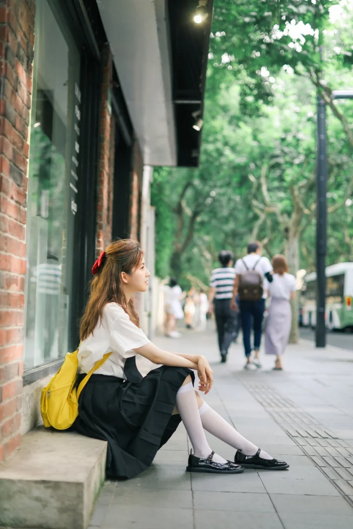 一名年轻女子坐在砖墙建筑旁的人行道台阶上，背景是树木和街道上的行人。