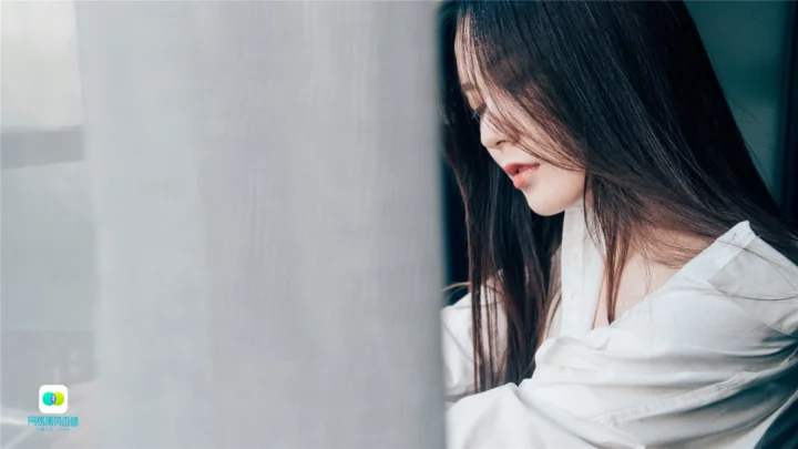 긴 머리의 여성이 흰 셔츠를 입고 창가에 앉아 왼쪽에서 부드러운 빛이 들어옵니다.